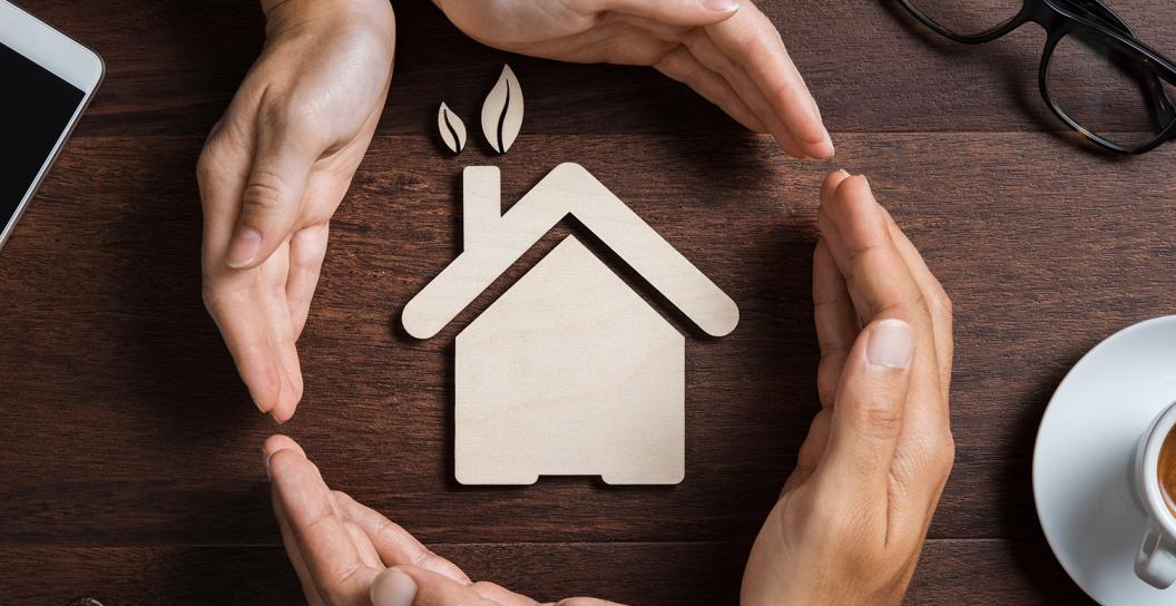 A paper cut out in the shape of a house is laying flat on a wooden table. Two sets of hands are forming a circle around the house.