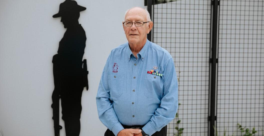 Elderly man wearing medal ribbons posing for camera standing next to Anzac sculpture