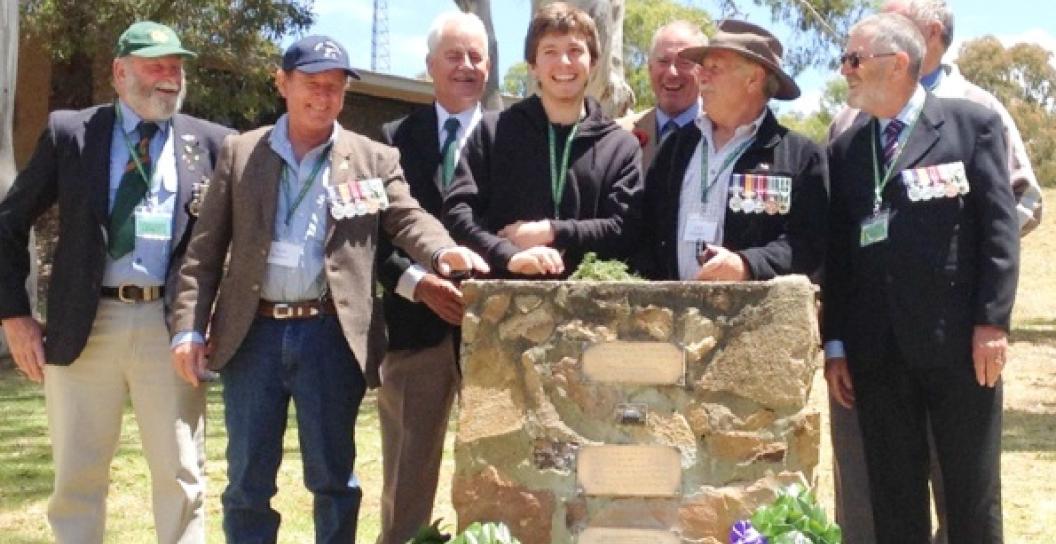 Young man at small memorial surrounded by mostly middle-aged or older men wearing medals