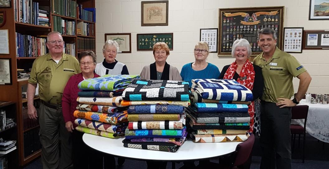 A group of people standing behind a table that holds folded patchwork quilts