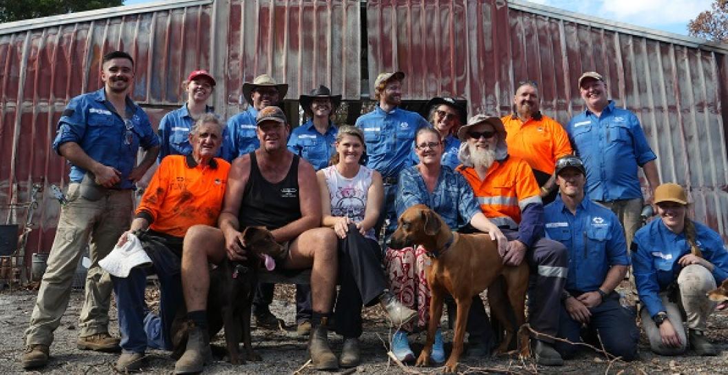 A group of people in hi-vis and working clothing stand in front of a tin shed. The front row is seated.