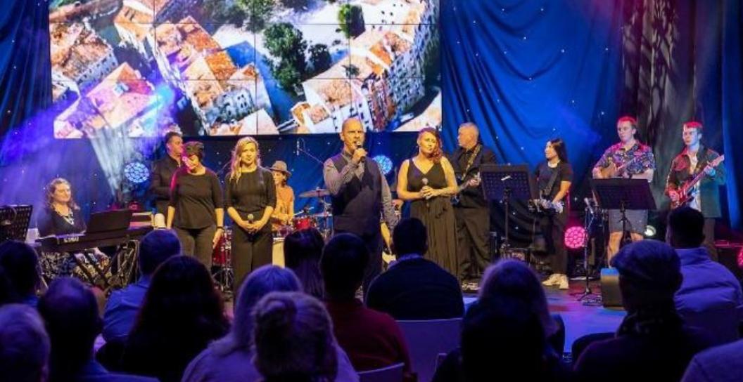 Several women on stage singing to audience, with musicians behind them.