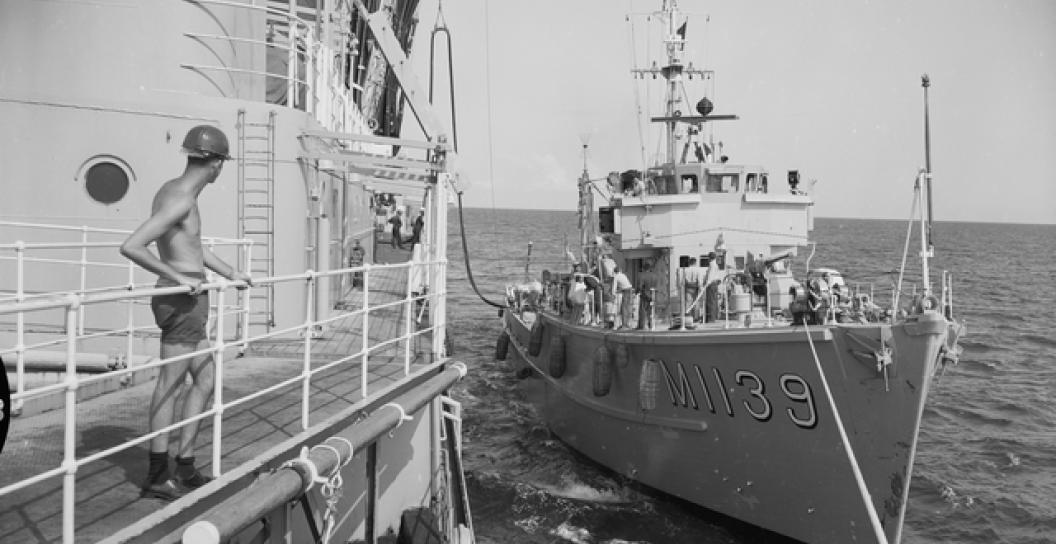 Small warship docks alongside larger Navy ship at sea, while a sailor looks on