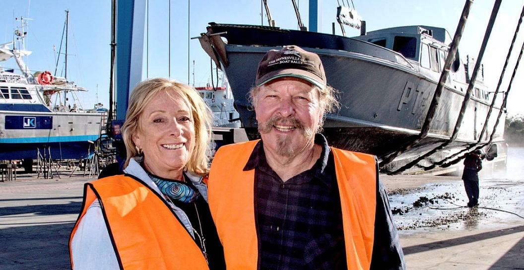 Couple pose for camera in front of grey boat suspended in harness
