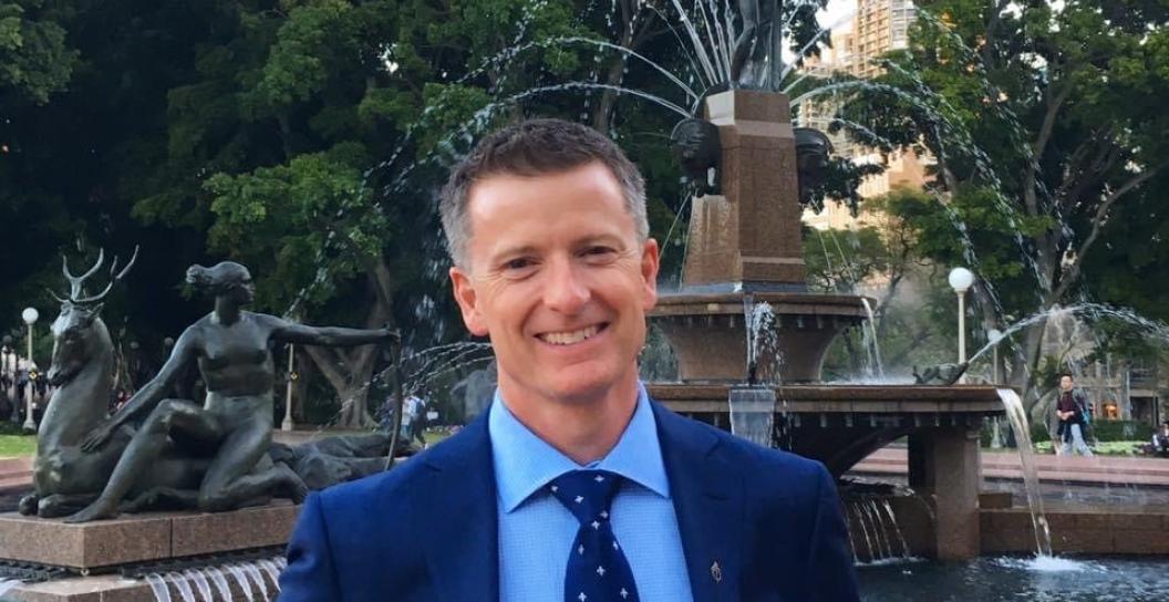 Man in suit smiling at camera with Sydney's Hyde Park fountain in background