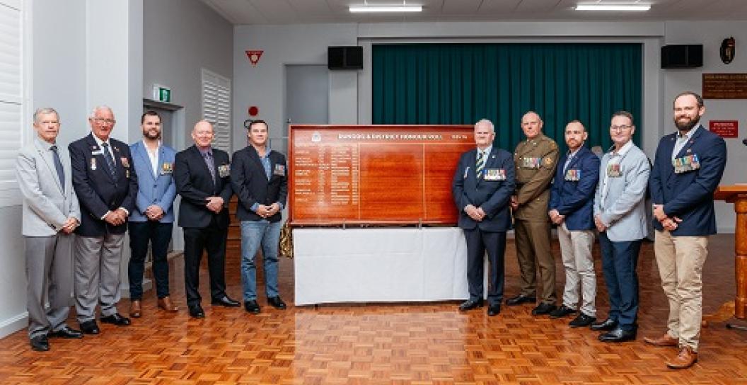 Ten men pose next to honour roll inside hall