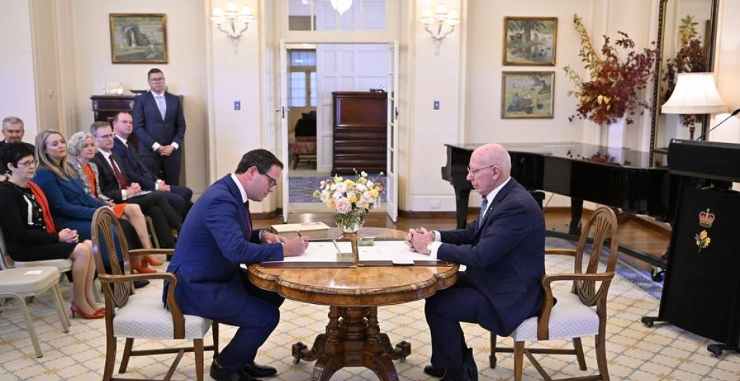 Man in suit writes on document on table with older man looking on and small audience watching