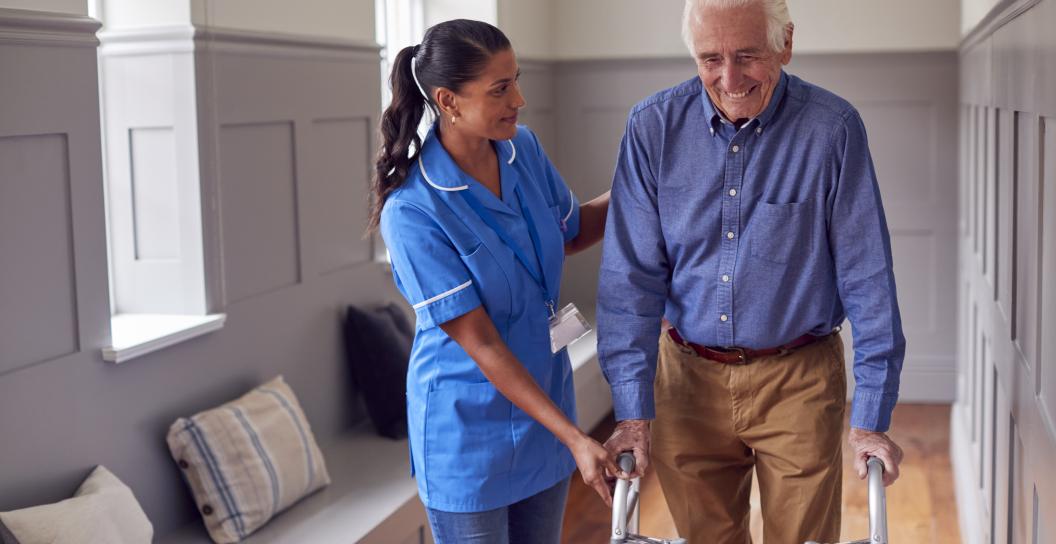 A senior man using a walking frame is being helped by a care worker