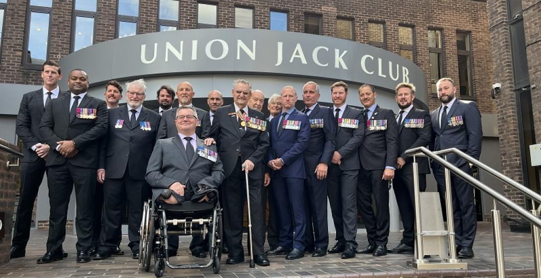 About 15 men in suits, one in a wheelchair, posing out the front of the Union Jack Club
