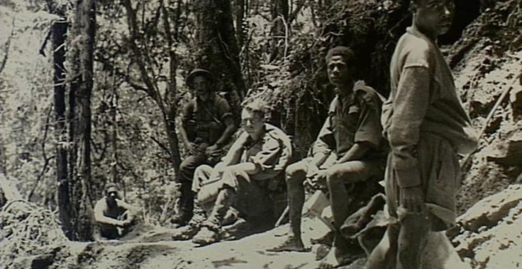 Black and white photo of Australian soldiers sitting next to Papuan bearers