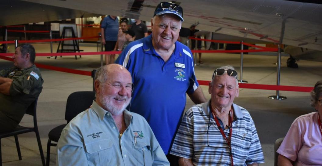 Three older men smiling, seated next to aircraft in hangar