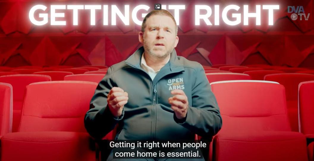 Video still showing man sitting on chair in auditorium wearing Open Arms top