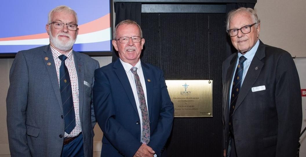 Three older men in suits pose smiling next to plaque