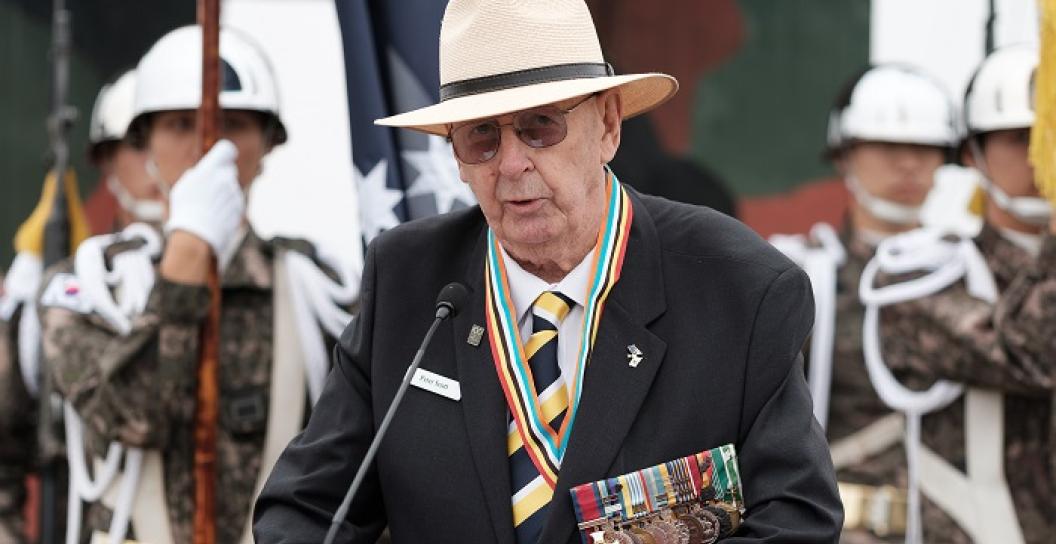Elderly man wearing medals speaking at lectern with Asian soldiers behind him