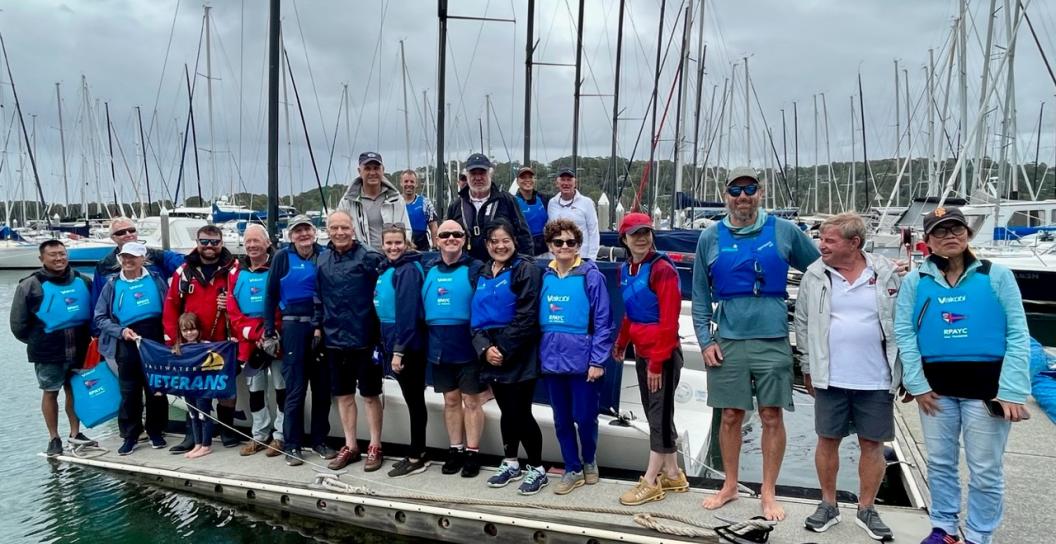 About 20 people stand on marina next to sailing boats