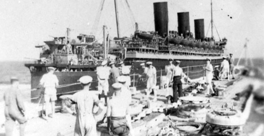 The Queen Mary, a British ocean liner, steams alongside the smaller Kent class cruiser HMAS Canberra, in the Atlantic during the Second World War. Some of the crew are on deck watching the Queen Mary sail past.