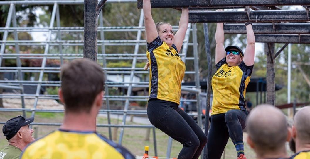Two people wearing Team Veteran T-shirts on obstacle course, watched by others in their team