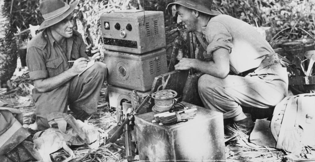Two men in Army uniform crouching by an old radio in the jungle