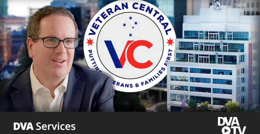 Minister Keogh wearing glasses smiling, with building in the background and a circle veterans centeral logo in the middle