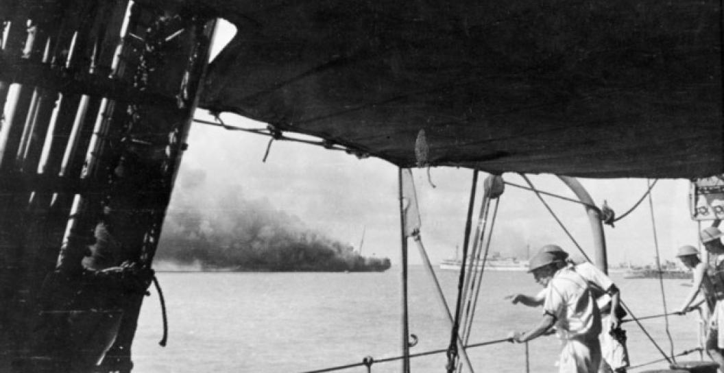 Crew members of the depot ship HMAS Platypus survey the harbour after the first Japanese air raid on Darwin, 19 February 1942. In the centre, the transport ship HMAT Zealandia is burning, and the hospital ship HMAS Manunda is to its right.