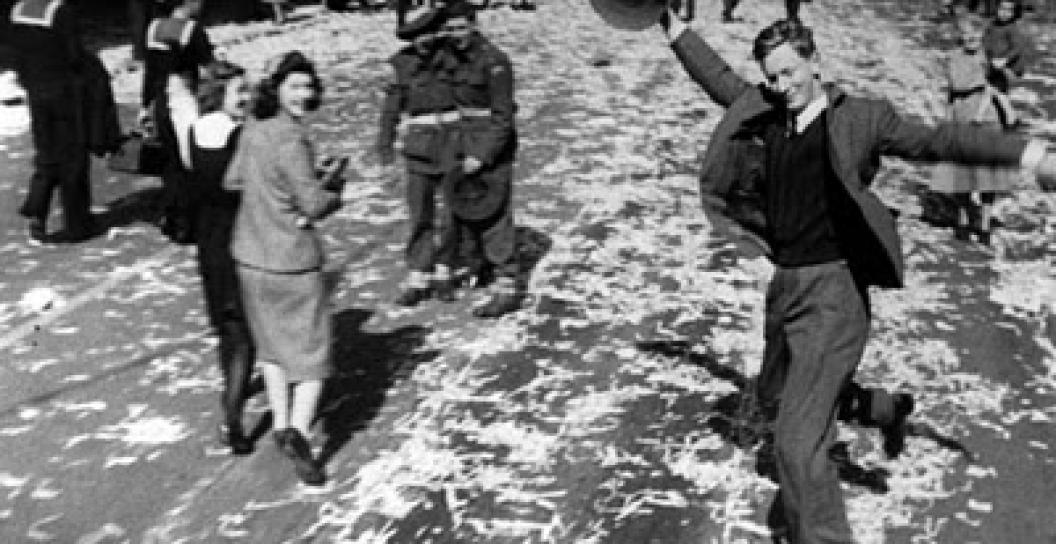 Black and white photo of man in suit dancing and smiling on busy street covered in ticker tape.
